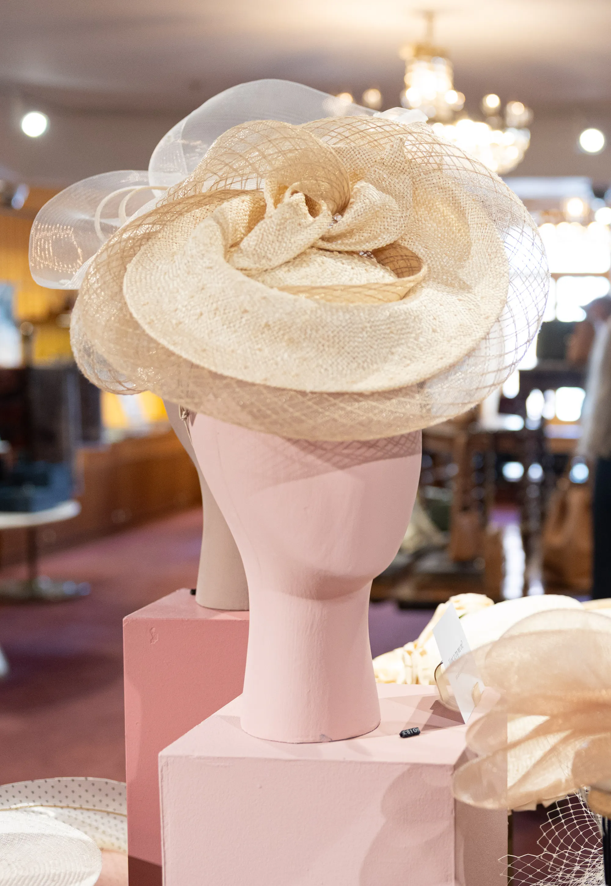 A beige sinamay fascinator featuring mesh loops and a wide brim, on display alongside other millinery hats.