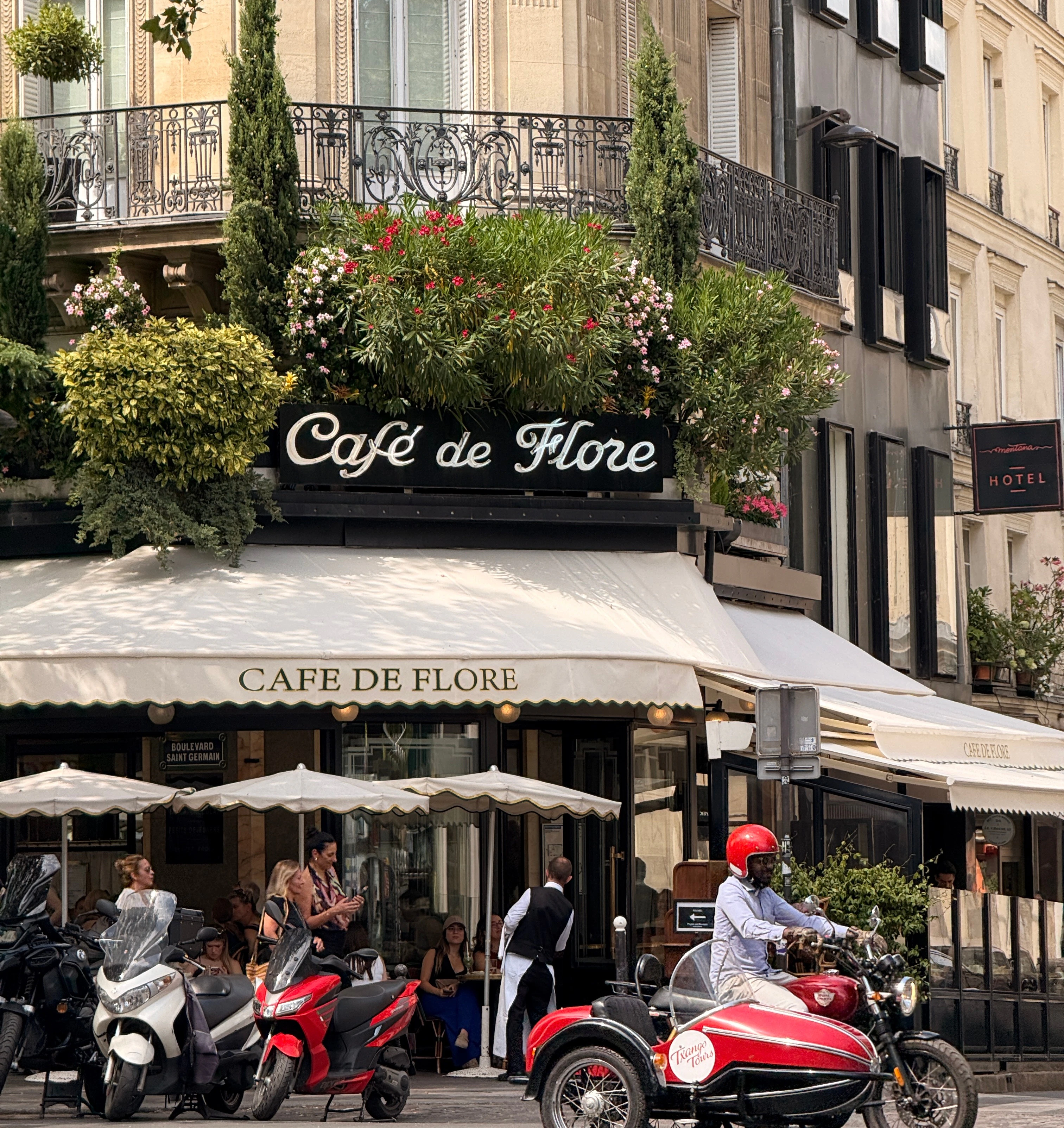 Paris street view of Café de Flore with overhanging flowers and a vintage sidecar parked in front.