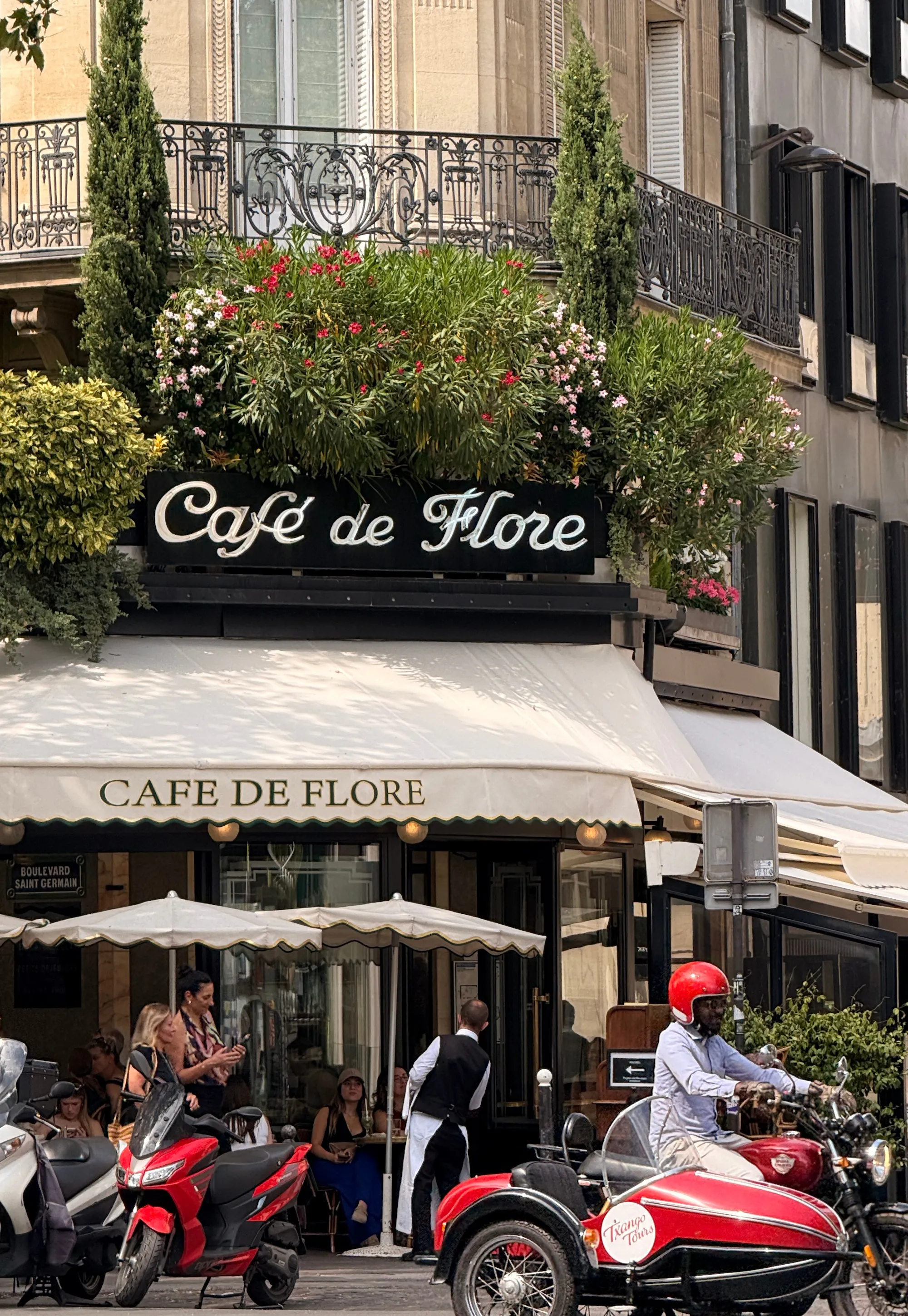 Paris street view of Café de Flore with overhanging flowers and a vintage sidecar parked in front.
