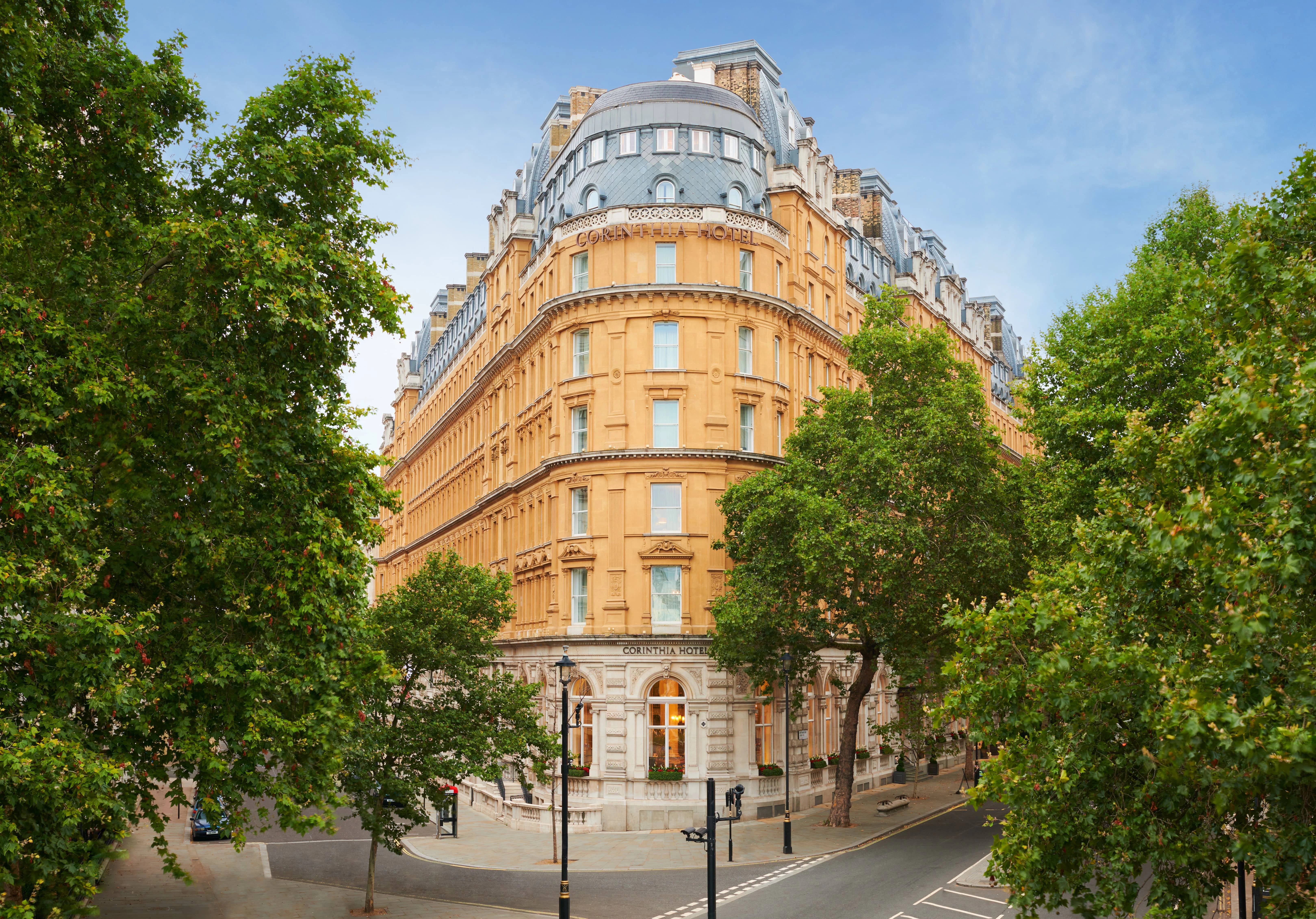 The curved, honey-stone facade of the Corinthia Hotel London, framed by plane trees on a clear day.