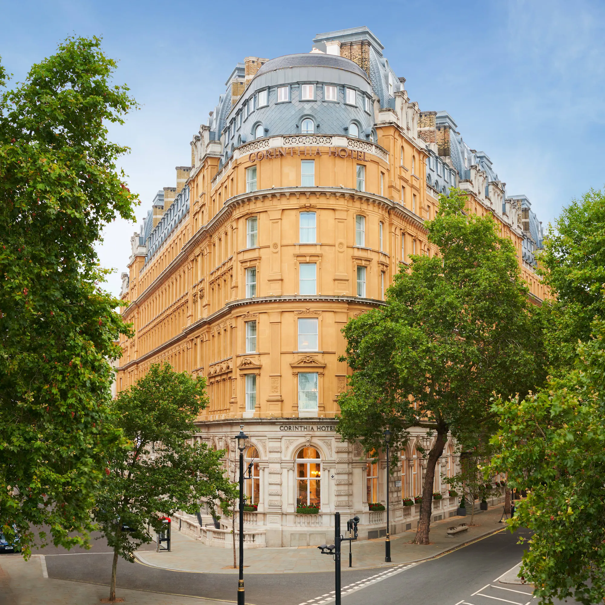 The curved, honey-stone facade of the Corinthia Hotel London, framed by plane trees on a clear day.