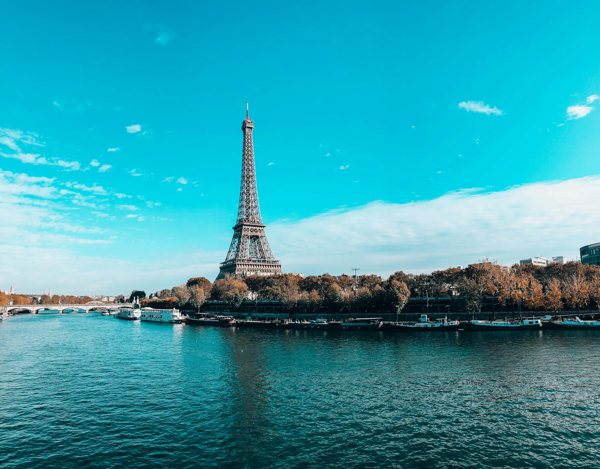 The Eiffel Tower rising above autumn trees, seen across the Seine lined with moored barges.