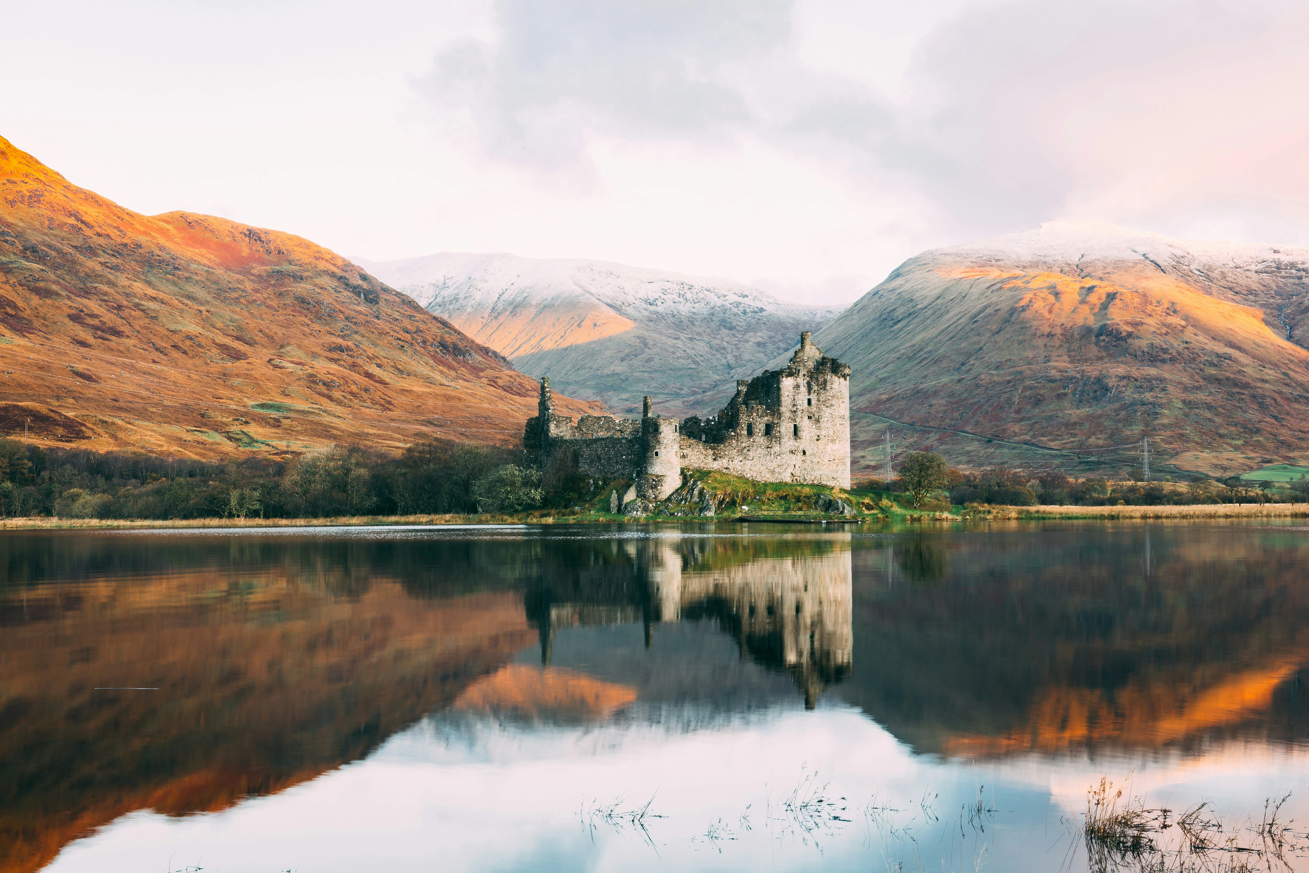 Kilchurn Castle's ruined towers reflected in Loch Awe, framed by autumn hills and snow-capped peaks.