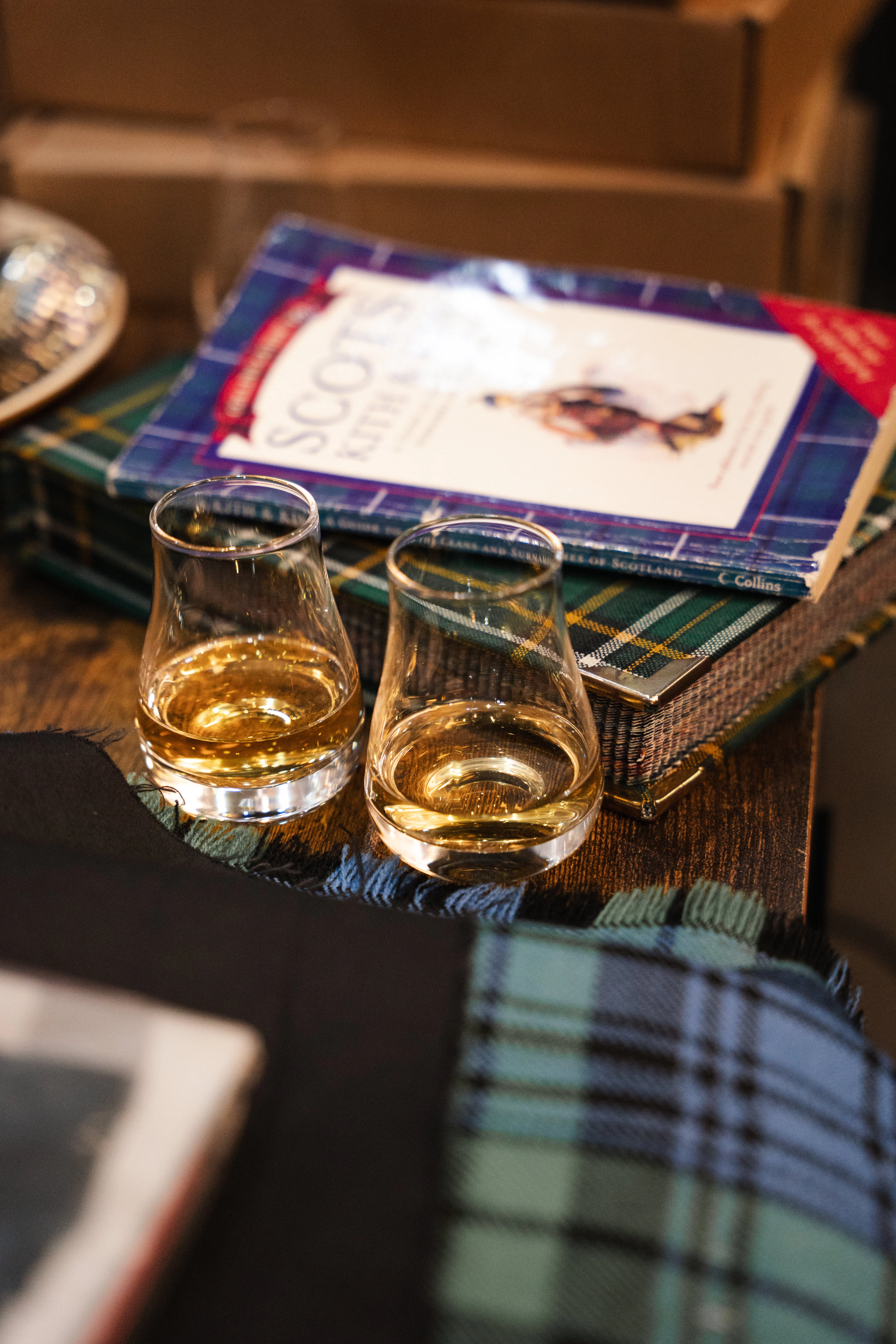 Two glasses of whisky beside a Scottish clan history book and tartan throw.