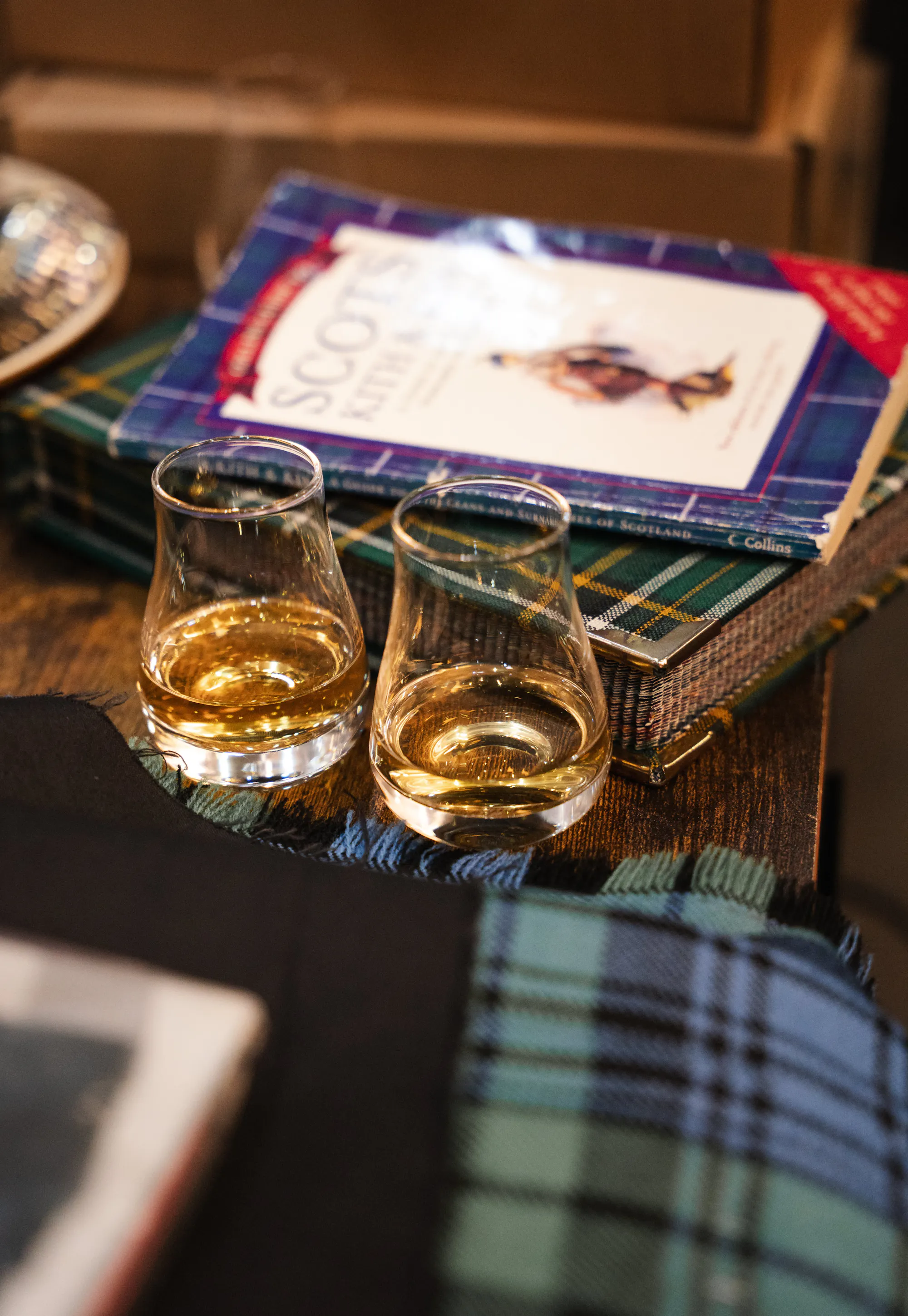 Two glasses of whisky beside a Scottish clan history book and tartan throw.