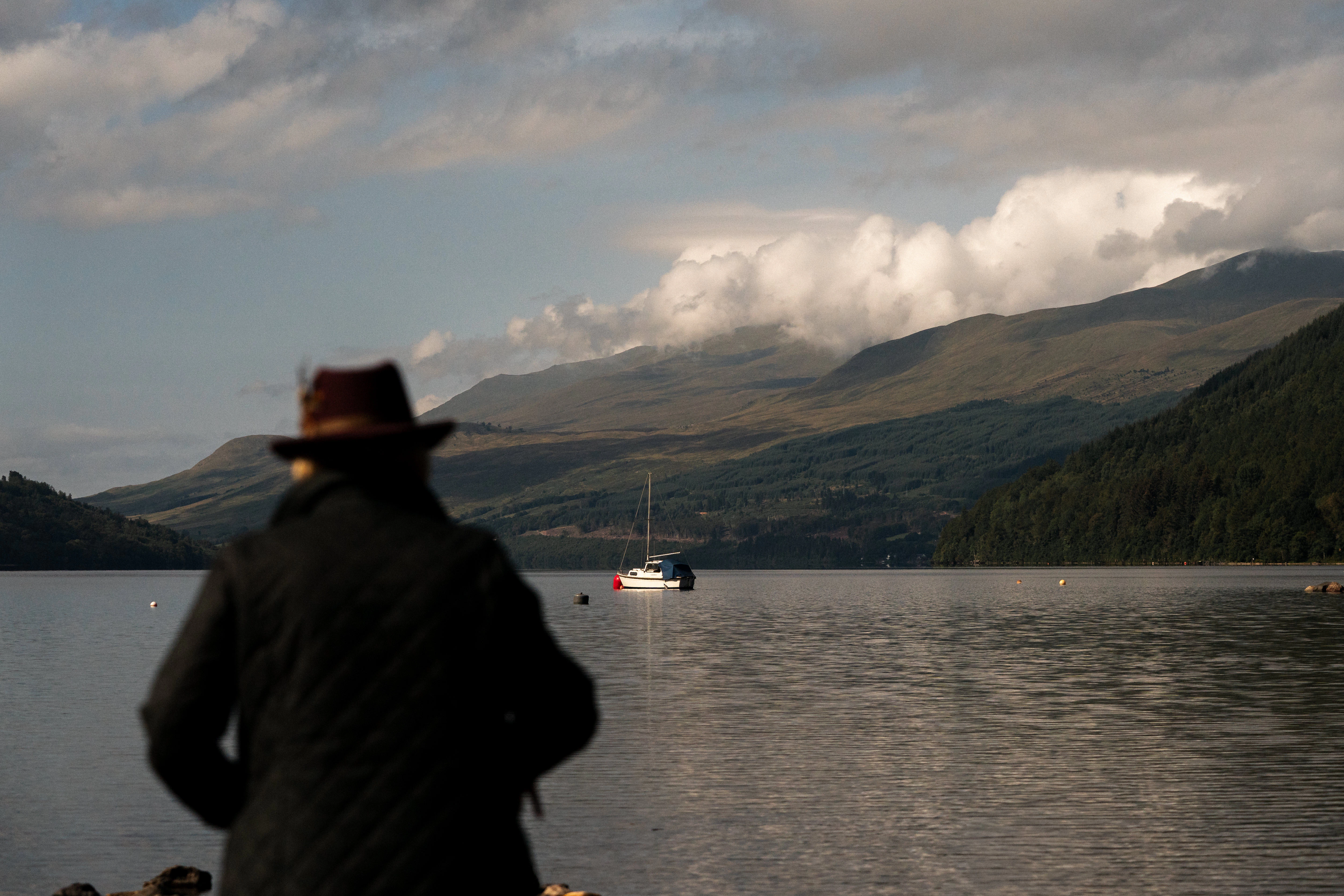 A traveller in a wide-brimmed hat watches a sailboat on a still Highland loch.