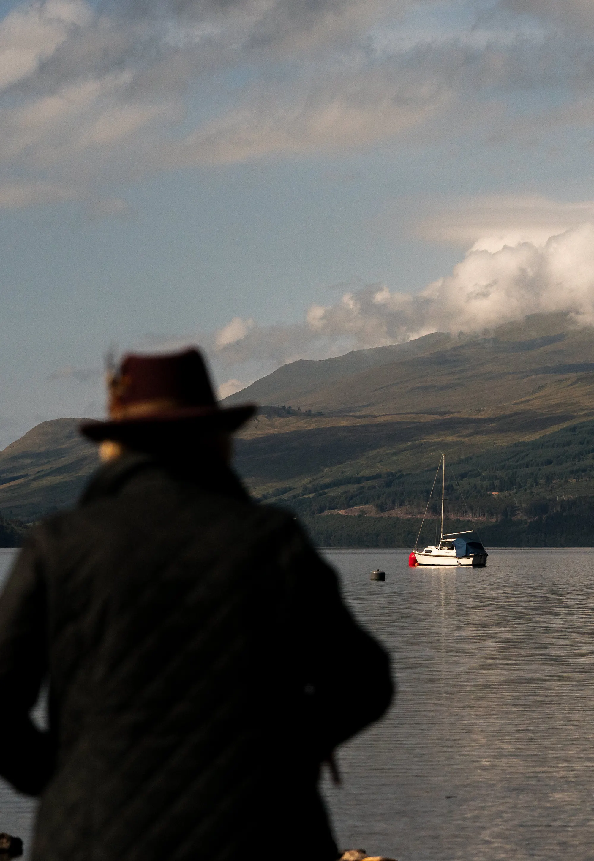 A traveller in a wide-brimmed hat watches a sailboat on a still Highland loch.