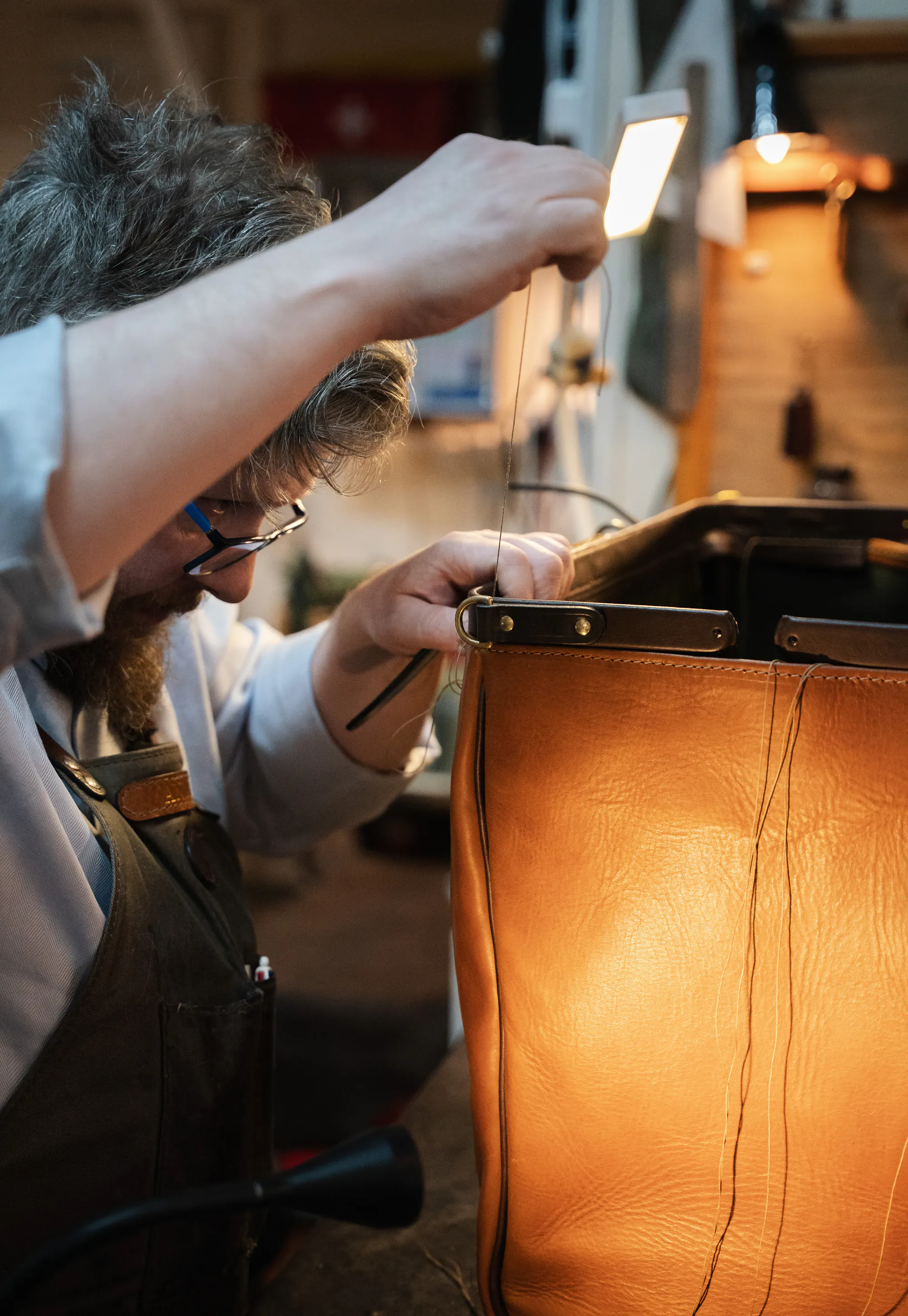 A craftsman hand-stitches a tan leather bag with waxed thread in his workshop.