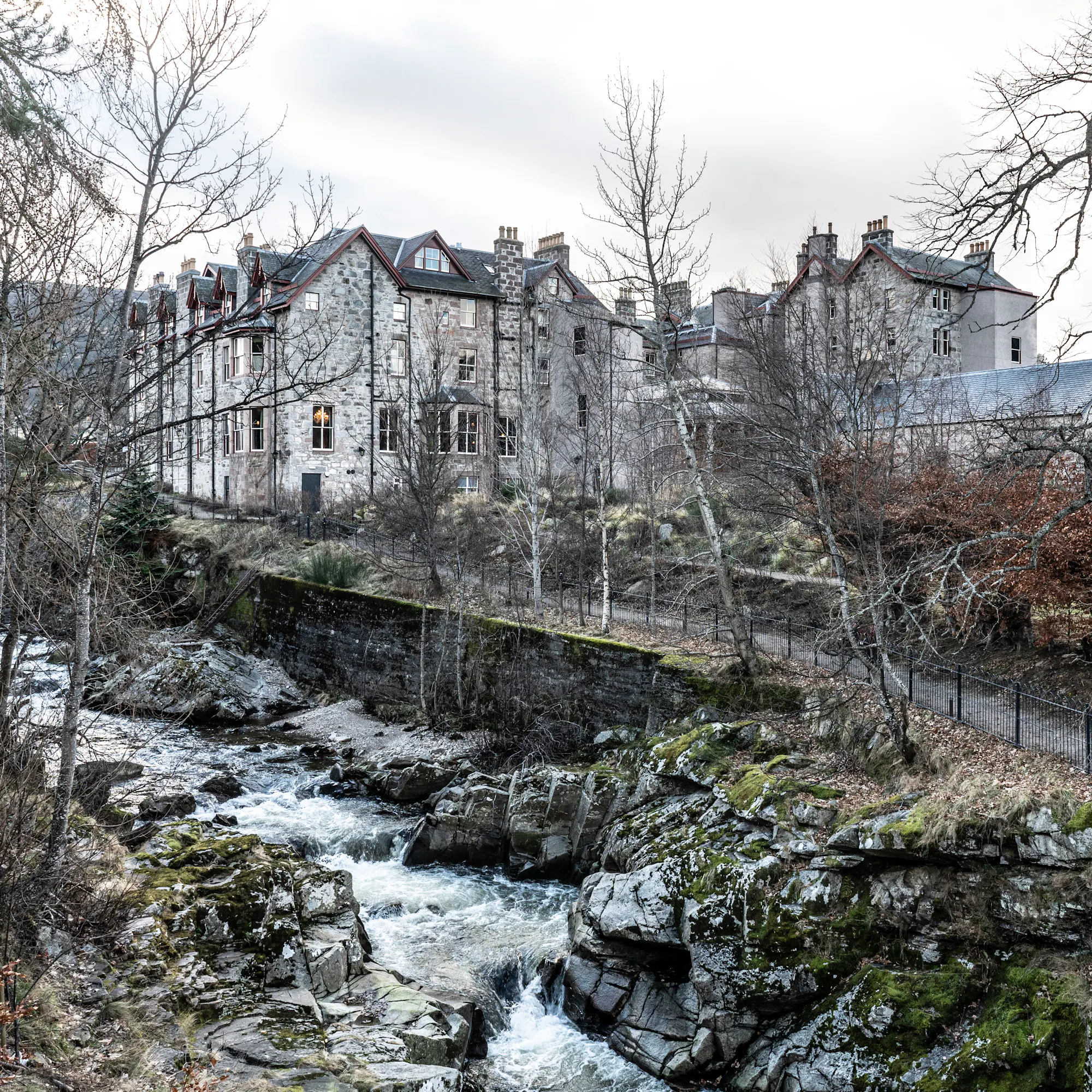 The Fife Arms hotel's stone facade above a rocky river bed, surrounded by bare winter trees.