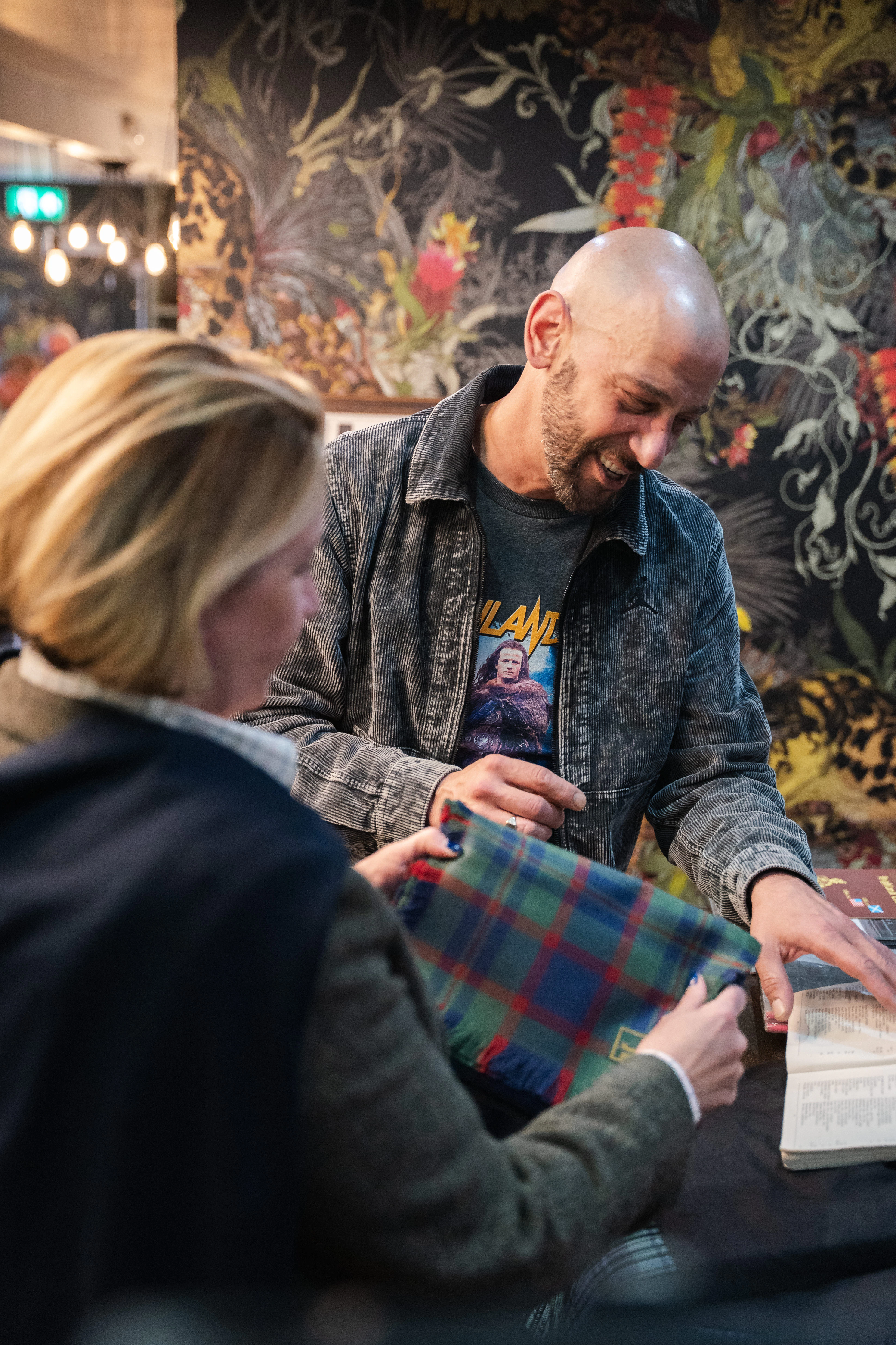 A man in a corduroy jacket laughs beside a woman holding a folded tartan, open clan book on the table in front of them.