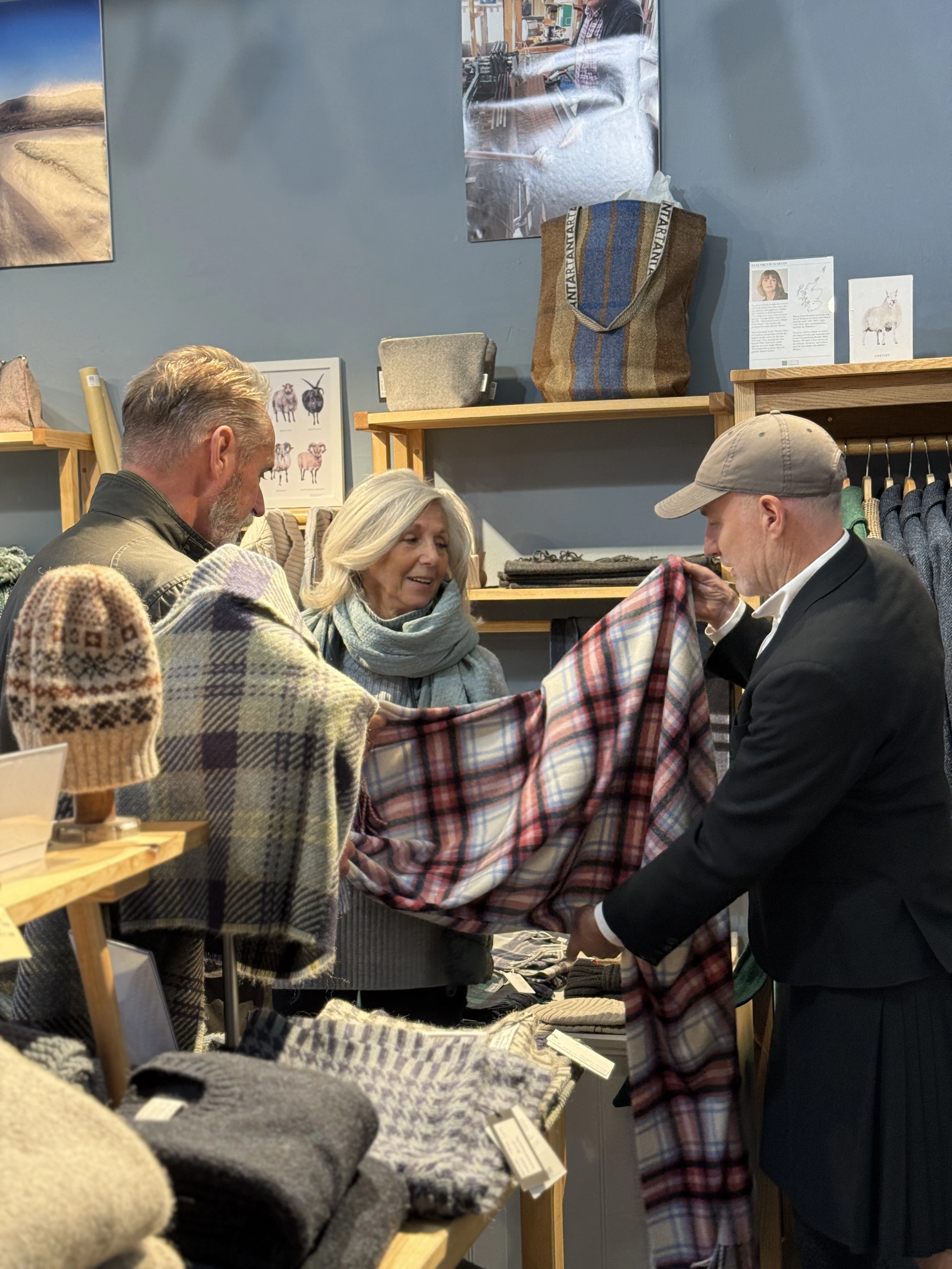 A man in a kilt holds up a tartan throw for a smiling couple in a woollen goods shop.